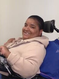 A black woman with very short, dark brown hair sits in a motorized wheelchair and is smiling at the camera.